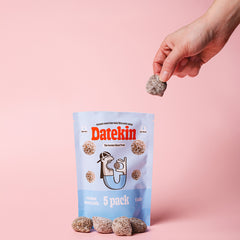Hand holding a Datekin coconut snack ball with packaging on a pink background