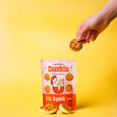 Hand holding a Datekin date snack cake above its packaging on a yellow background